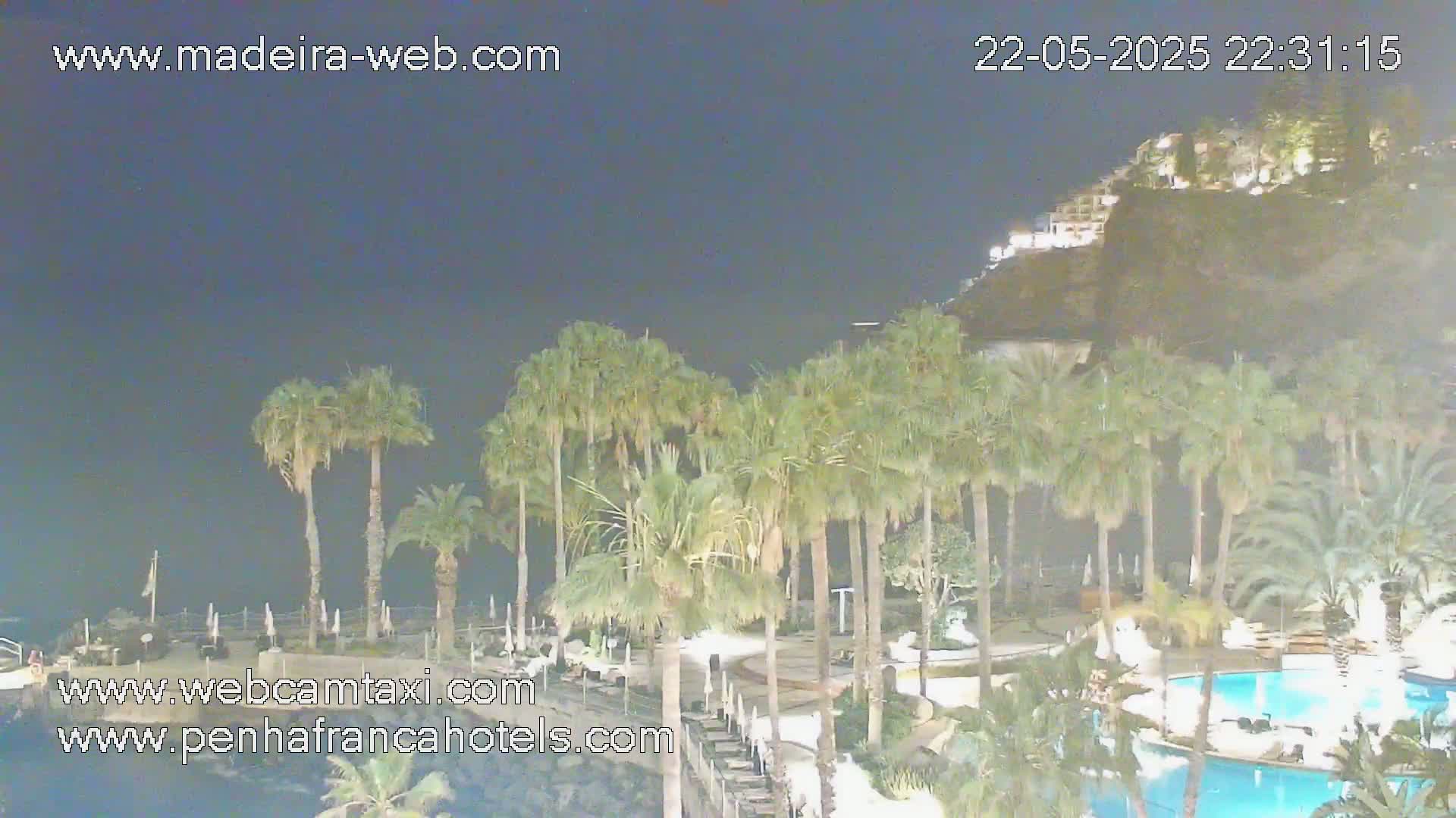 A nighttime view of a resort area with palm trees surrounding pools and buildings, situated on a cliffside overlooking the ocean under a clear, dark sky.