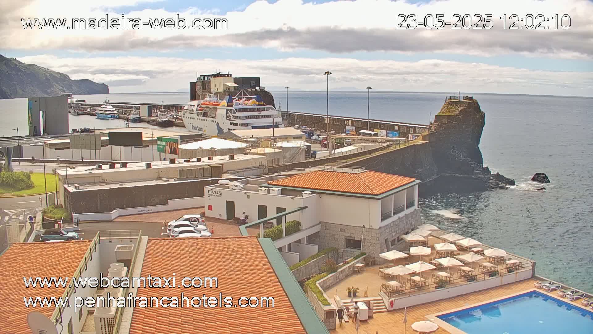 A partly cloudy day overlooks a harbor with a ferry docked, buildings, a swimming pool, and a rocky coastline.