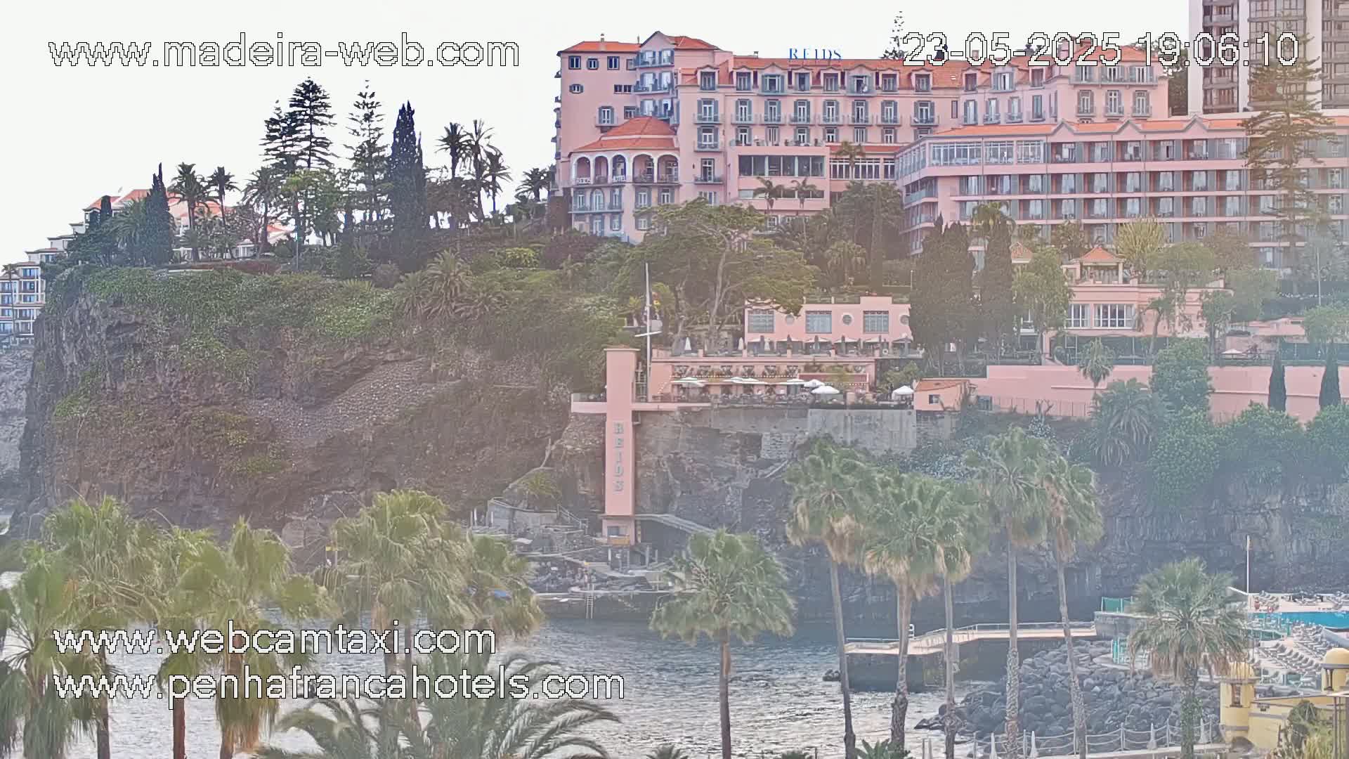 A large pink hotel complex sits atop a rocky cliff overlooking a calm body of water, surrounded by lush vegetation including palm trees under an overcast sky.