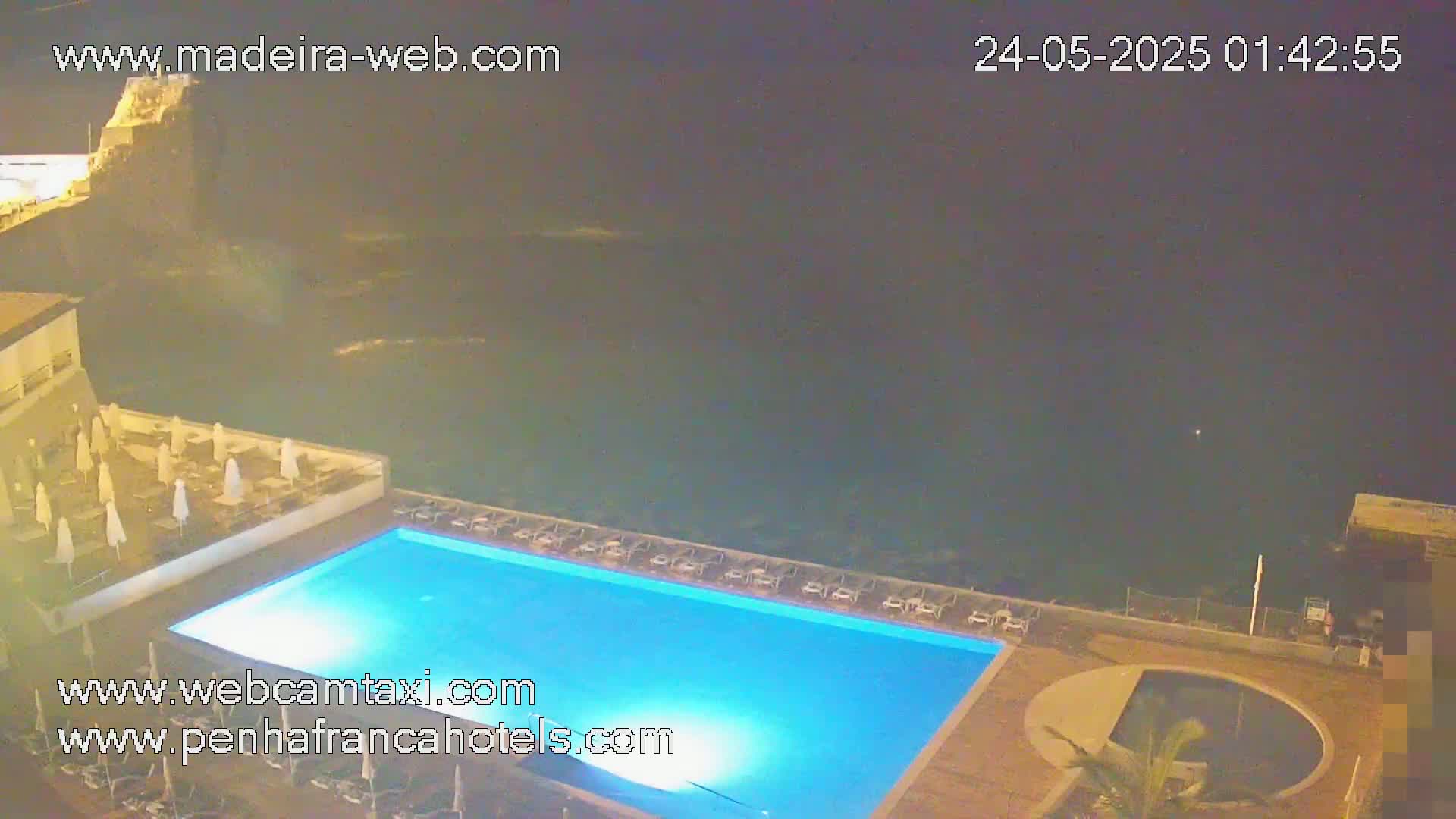 A nighttime aerial view shows a swimming pool and surrounding deck area at a resort overlooking a dark ocean.