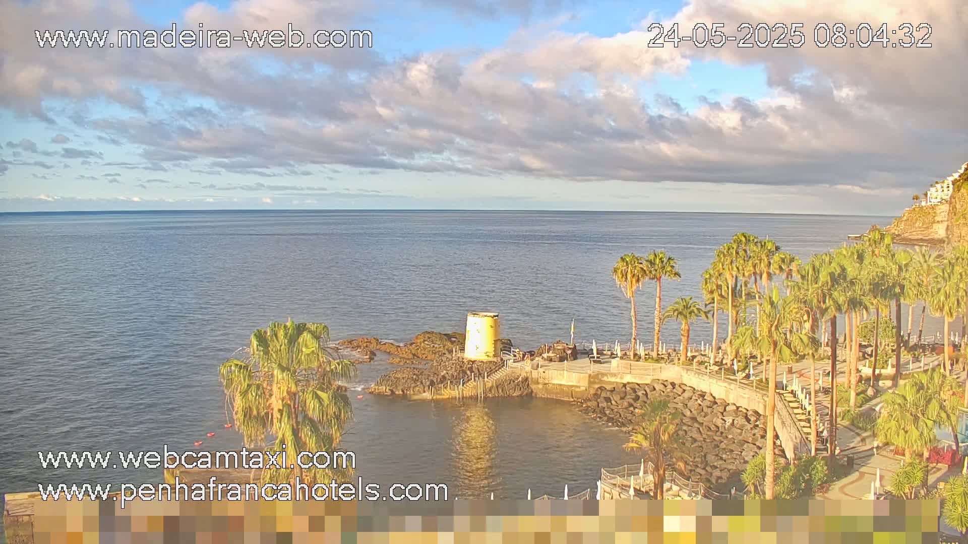 A partly cloudy day reveals a tranquil ocean scene with palm trees lining a rocky coast, featuring a small yellow structure near the water's edge.