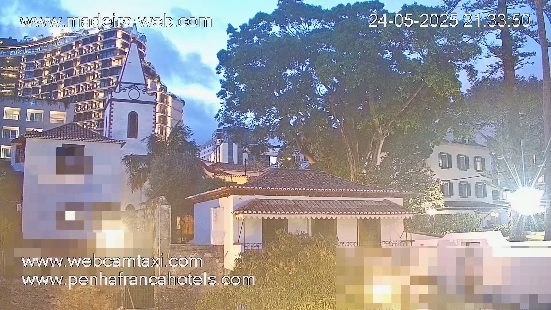 A small, white house with a red tile roof is nestled among larger buildings and lush greenery under a twilight sky.