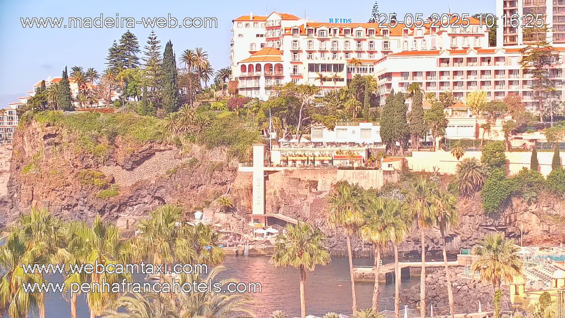 A seaside cliffside resort with numerous buildings, lush vegetation, and palm trees, under sunny skies.