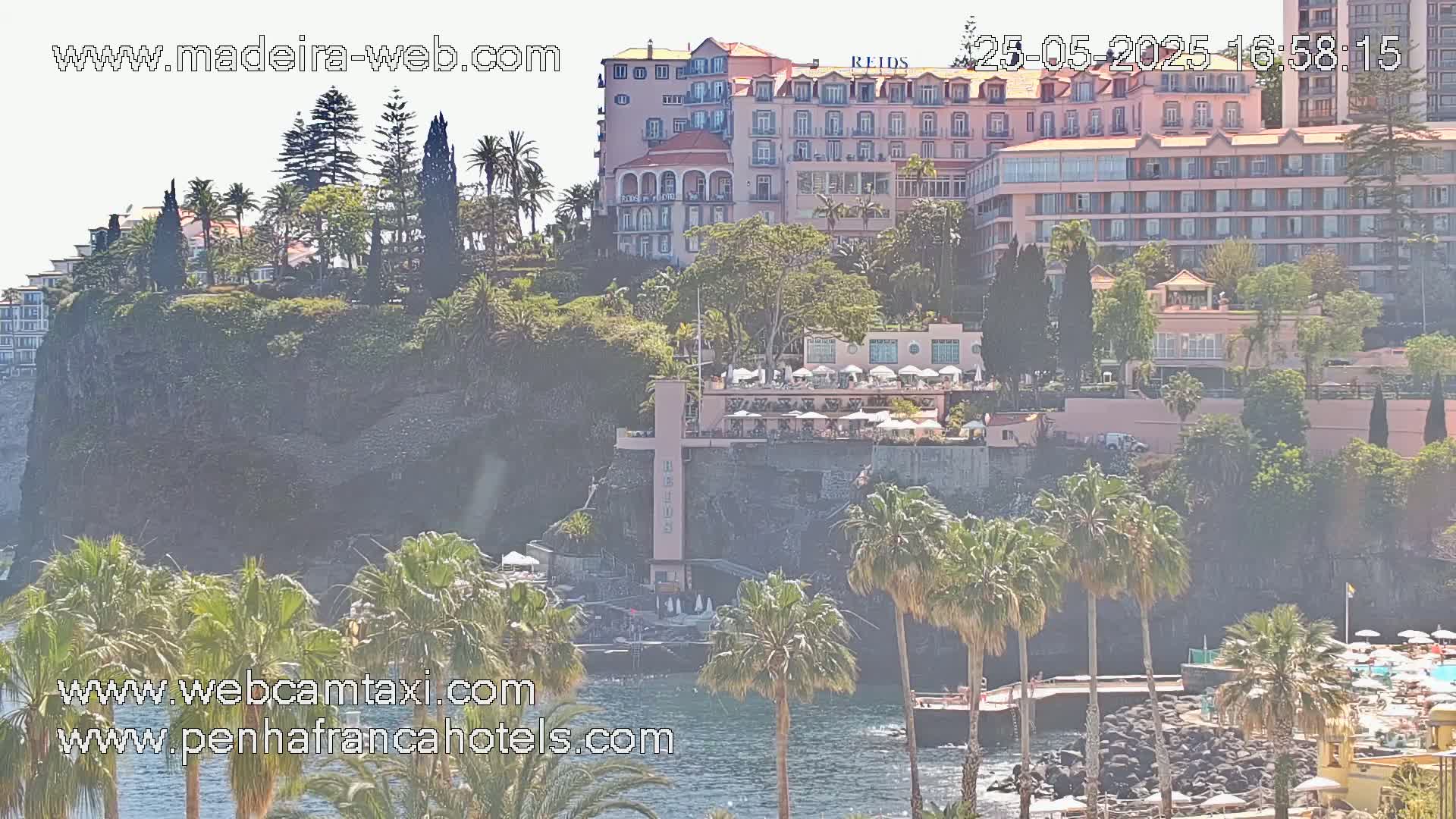 A seaside resort, situated on a cliff overlooking calm water, is partially obscured by hazy atmospheric conditions and lush vegetation, including palm trees.
