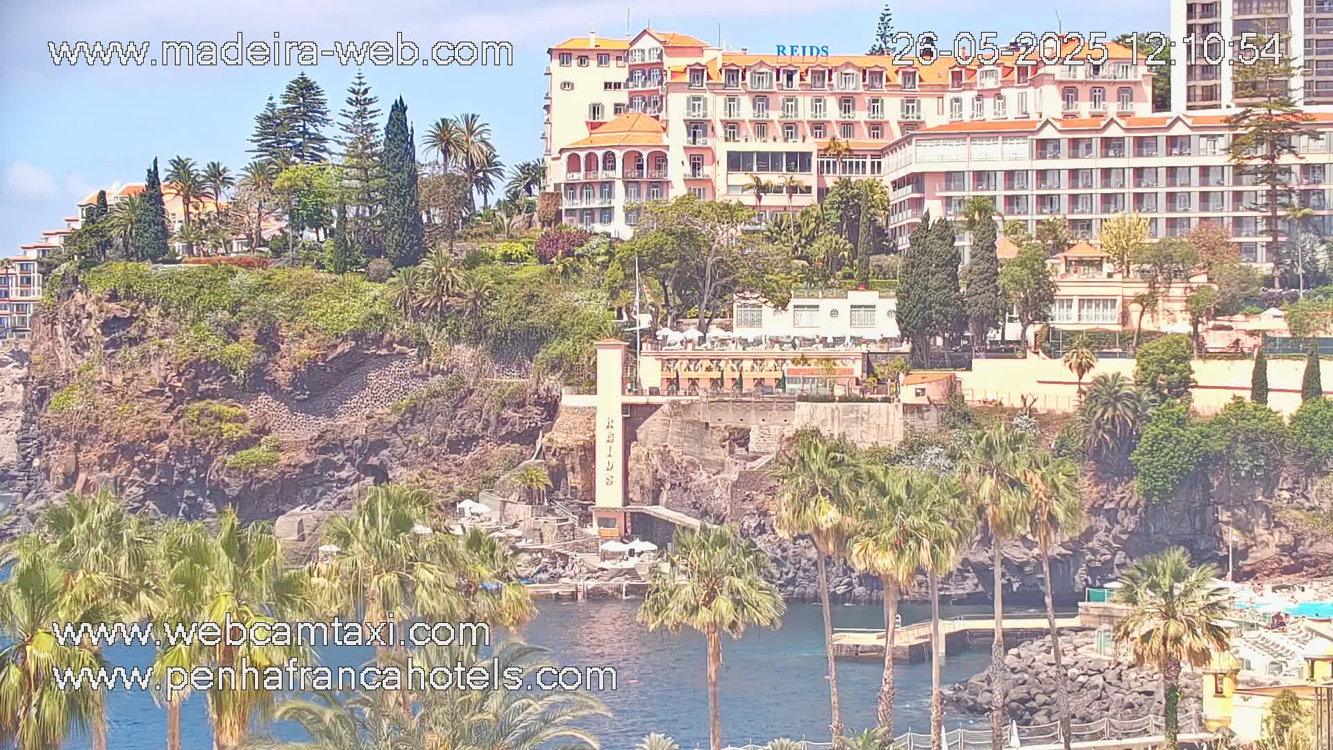 A coastal scene on a sunny day shows a large hotel perched on a cliff overlooking a calm body of water, surrounded by lush vegetation and palm trees.