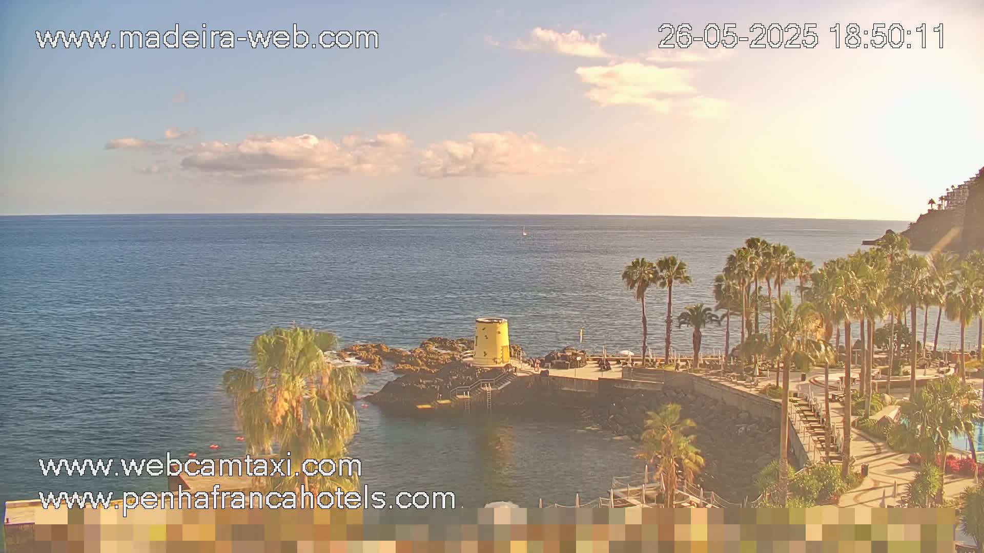 A partly cloudy sunset illuminates a rocky coastline with palm trees and a small yellow structure near calm ocean water.