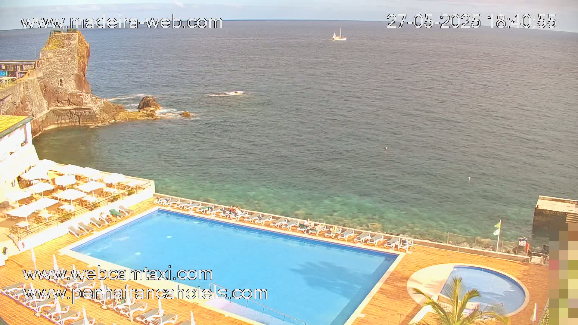 An outdoor swimming pool and small circular pool are situated on a hotel patio overlooking a calm ocean and a rocky outcrop with a stone tower on a sunny day.