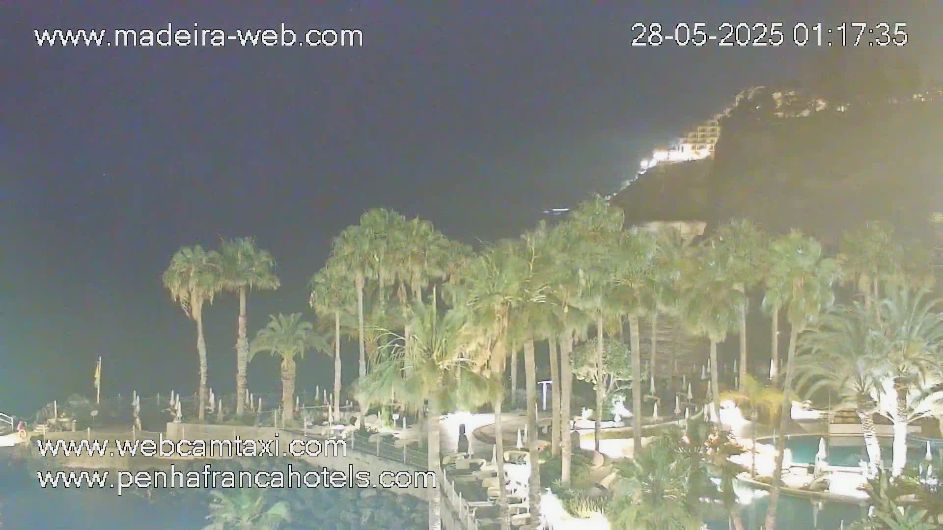 A nighttime view of a resort area with palm trees, pools, and a hillside with buildings illuminated in the background under a dark sky.