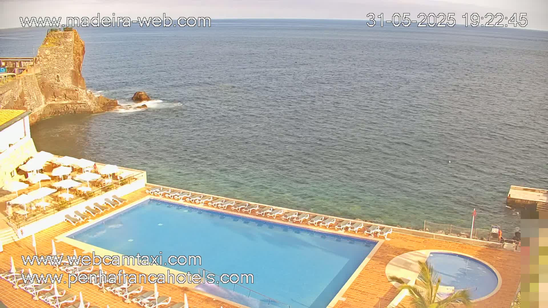 An outdoor pool area with lounge chairs and umbrellas overlooks a calm ocean next to a rocky cliff.