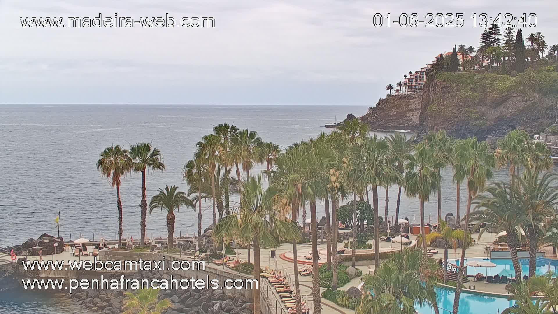A resort with palm trees and swimming pools overlooks a calm ocean under an overcast sky.