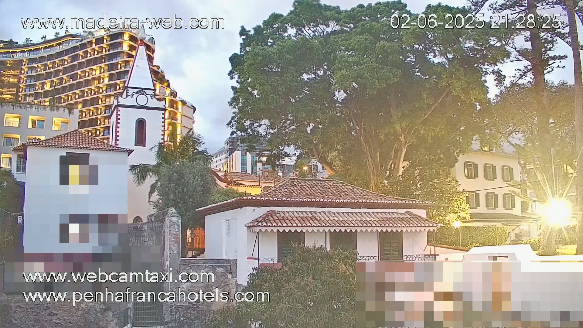 A white building with a red-tiled roof sits beside a church and a large hotel under a twilight sky.