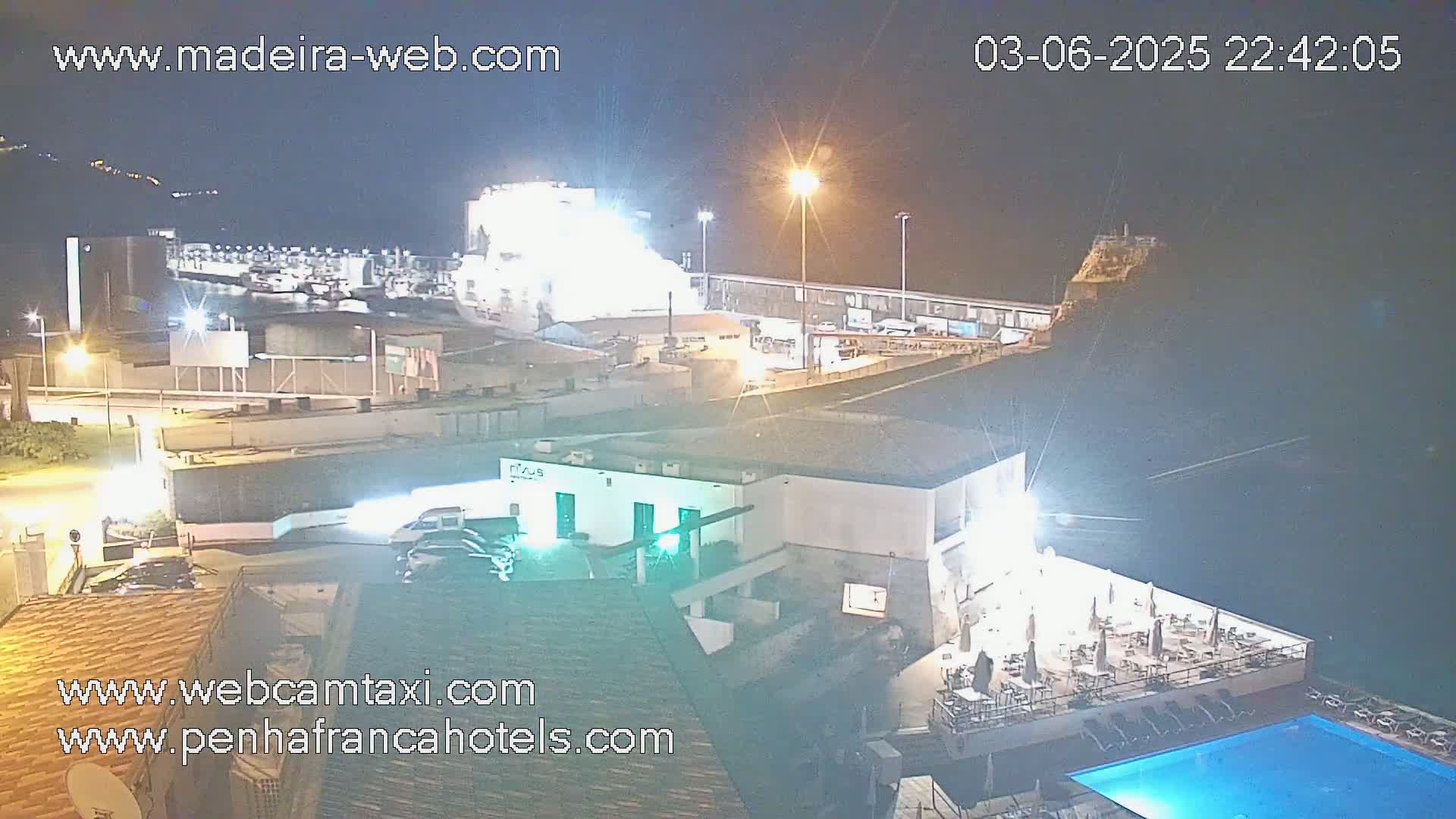 A nighttime aerial view of a harbor with a large ship docked, illuminated buildings, and a swimming pool.