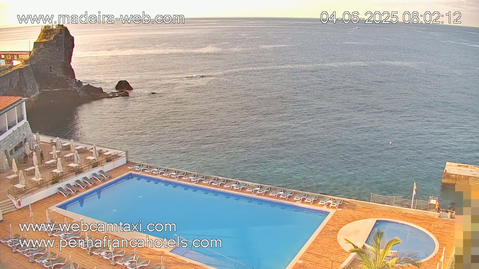 An outdoor pool and patio area at a hotel overlook a calm ocean on a partly sunny morning.