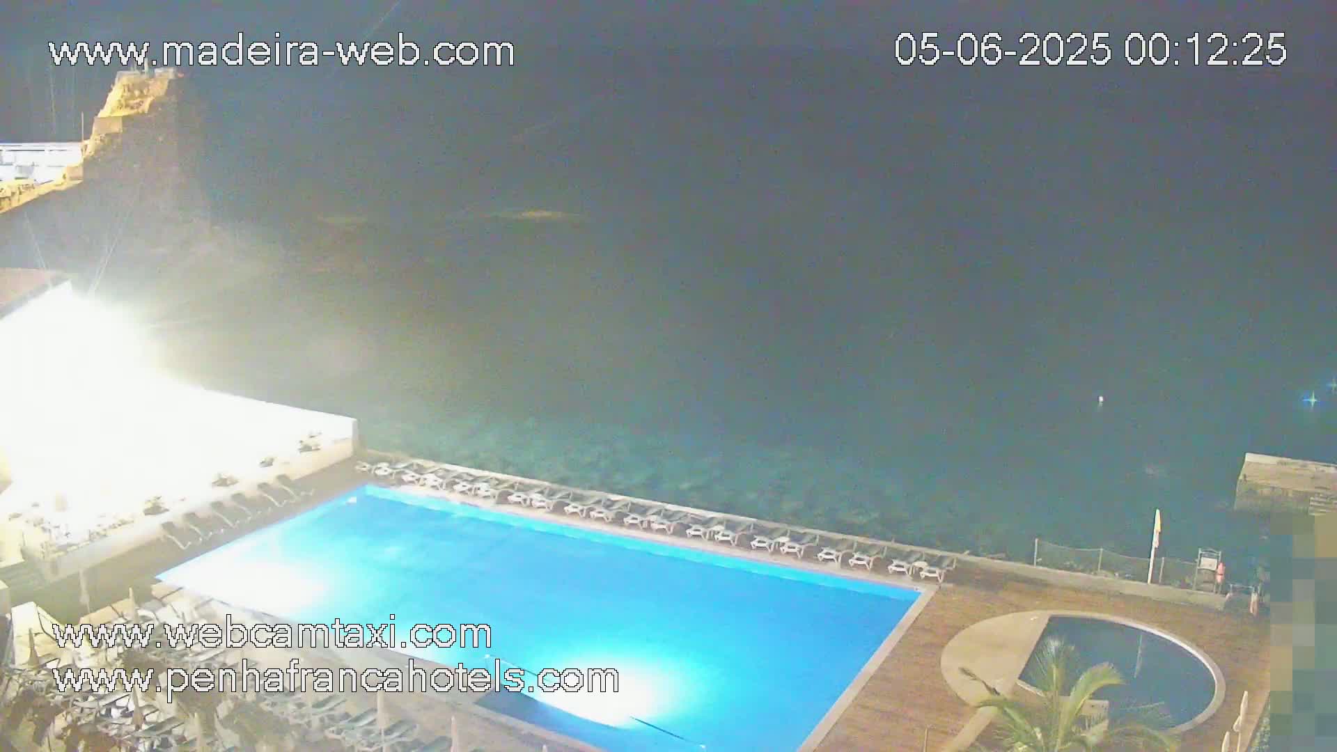 A nighttime view of a rectangular swimming pool next to a dark ocean, with lounge chairs surrounding the pool and a smaller round pool nearby.