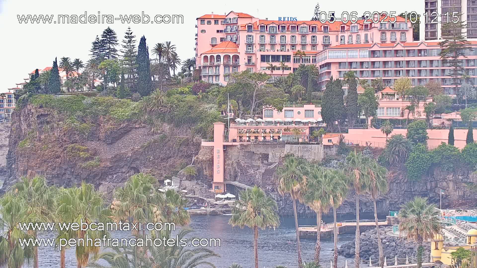 A large pink hotel complex sits on a cliff overlooking a calm body of water, surrounded by lush vegetation and palm trees under an overcast sky.