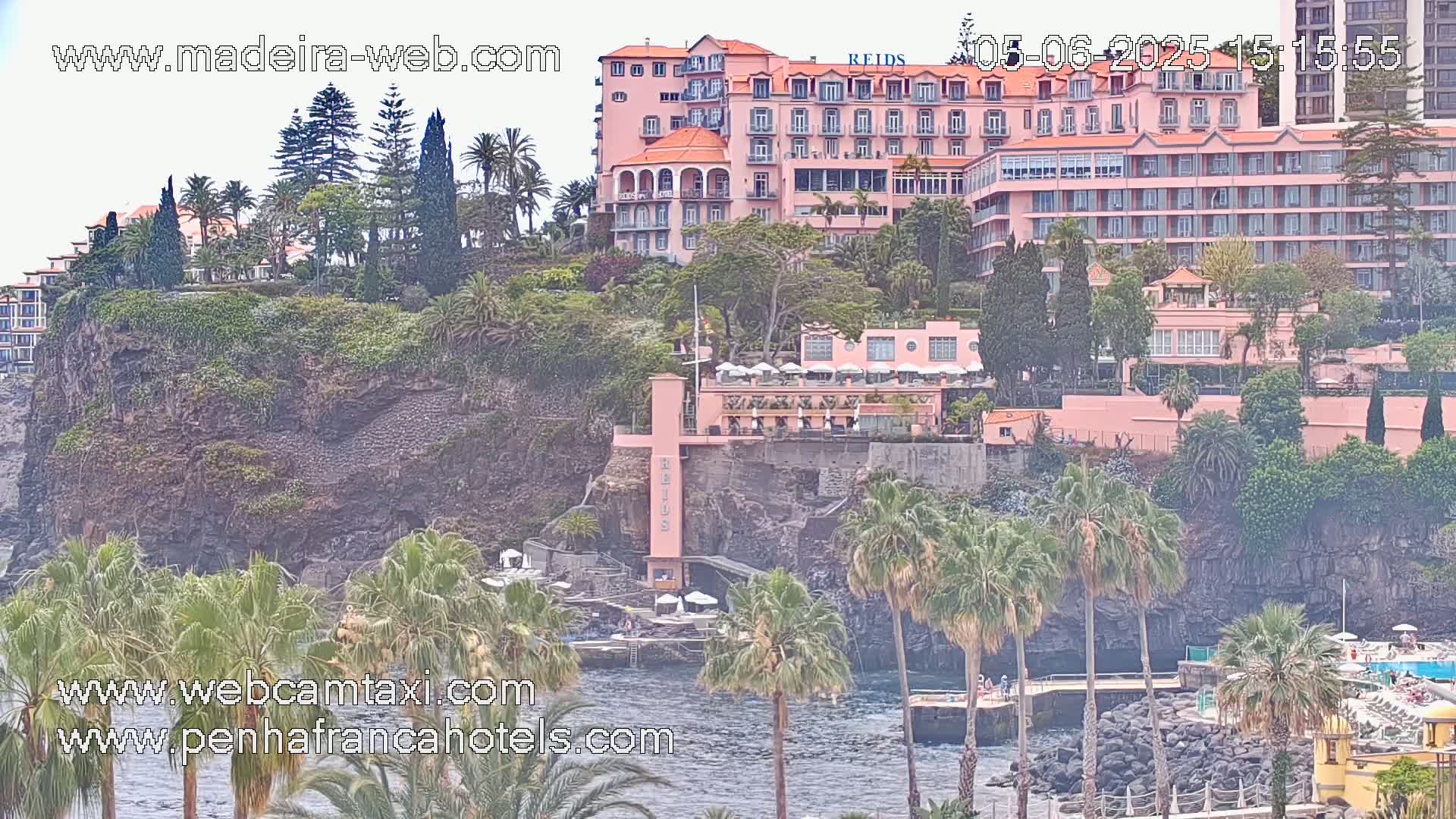 A pink hotel complex sits atop a rocky cliff overlooking a calm ocean, surrounded by lush vegetation and palm trees in hazy weather.