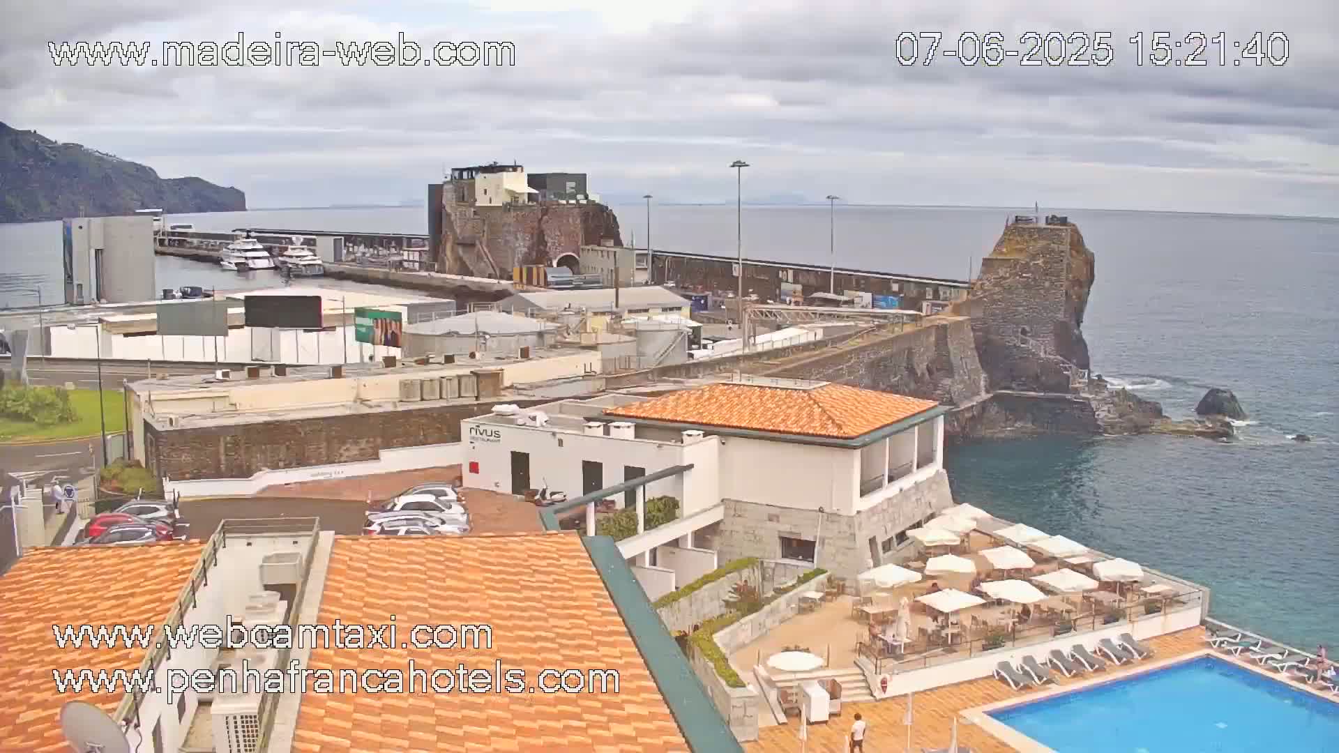 An overcast day shows a harbor with boats, buildings, and a hotel with an outdoor pool and patio area, all situated on a rocky coast.