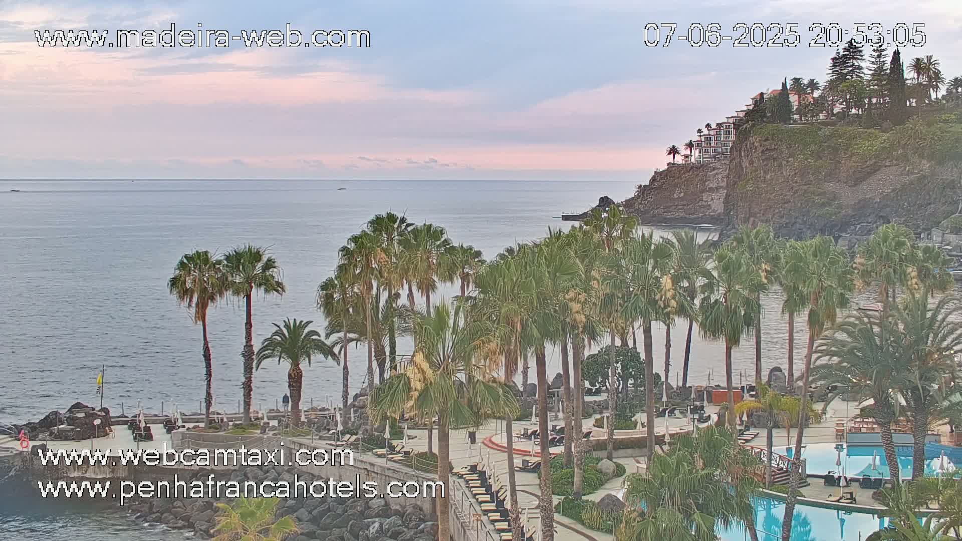 An ocean view from a resort shows a calm sea under a pale pink and gray sky, with palm trees surrounding a pool area and buildings on a cliff in the background.