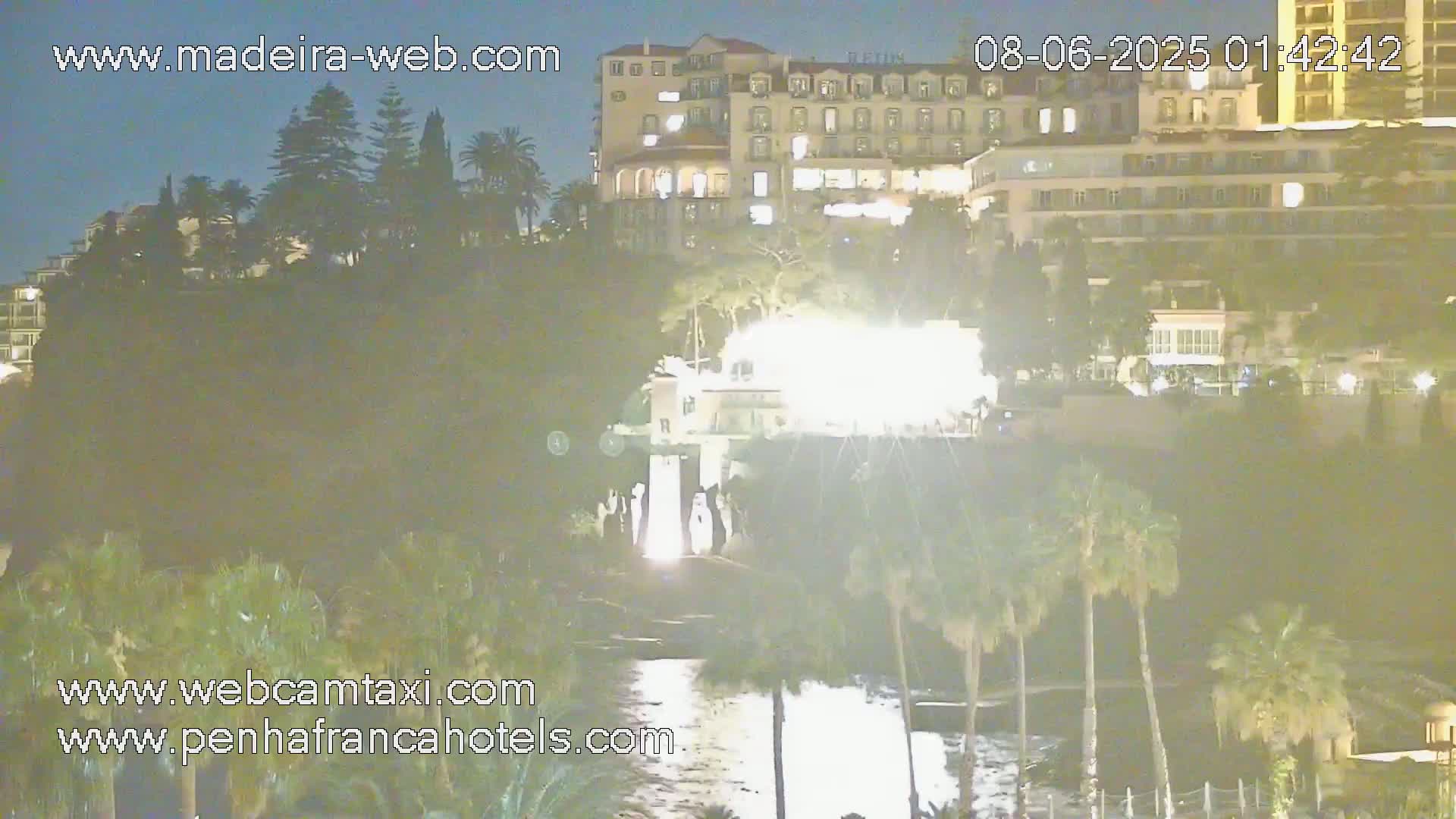 A nighttime view of a cliffside with a brightly lit building near the water, illuminated by artificial light, with palm trees and other vegetation.