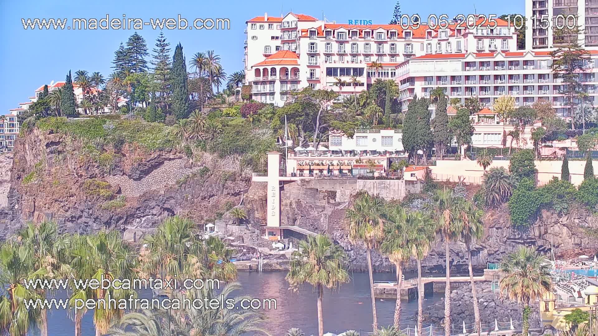 A cliffside view of hotels and buildings, lush vegetation including palm trees, and dark-colored volcanic rock formations overlooking a calm body of water under a sunny sky.
