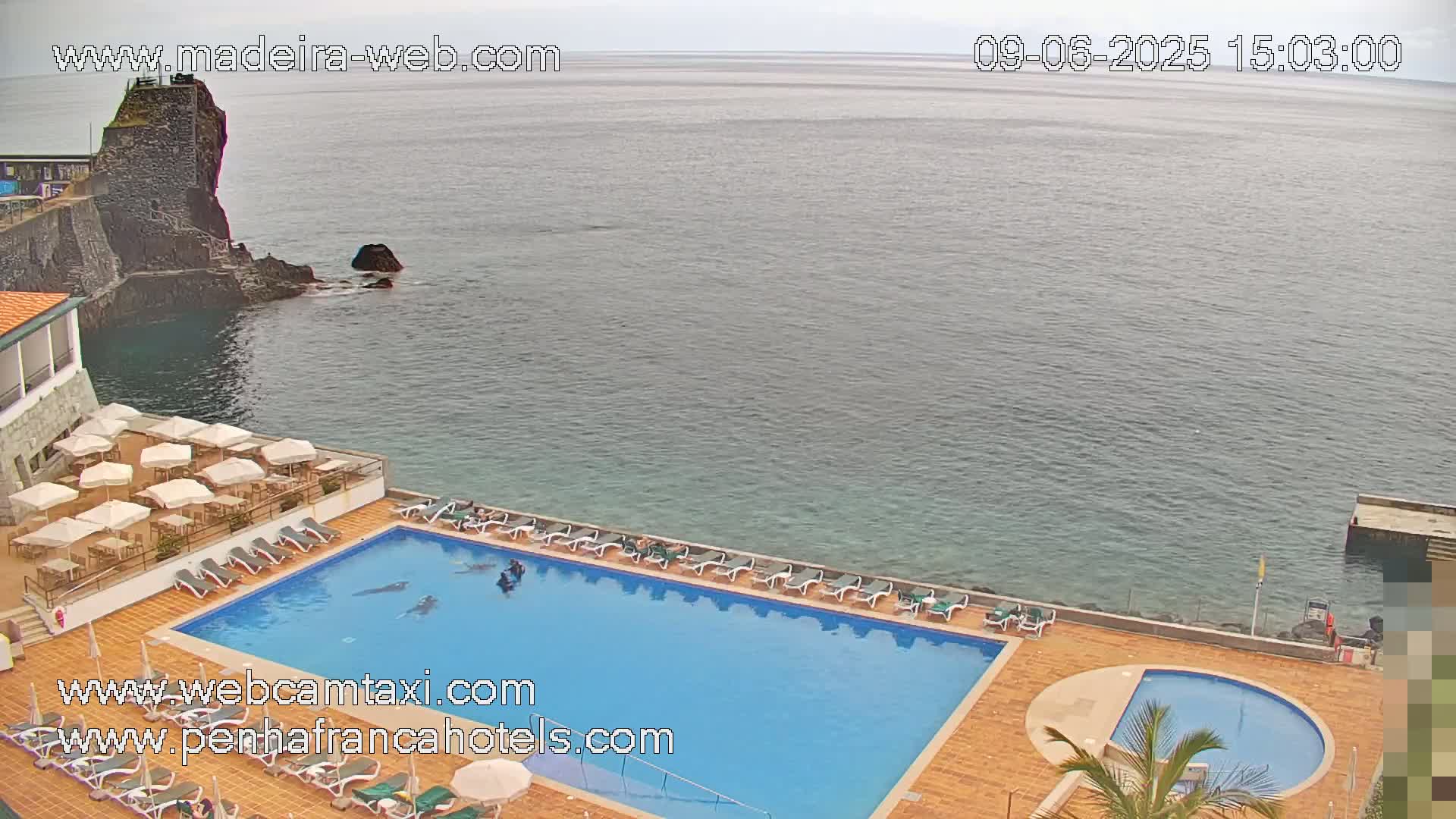 An outdoor pool with lounge chairs and umbrellas is situated on a cliff overlooking a calm ocean, with a stone structure visible in the water.