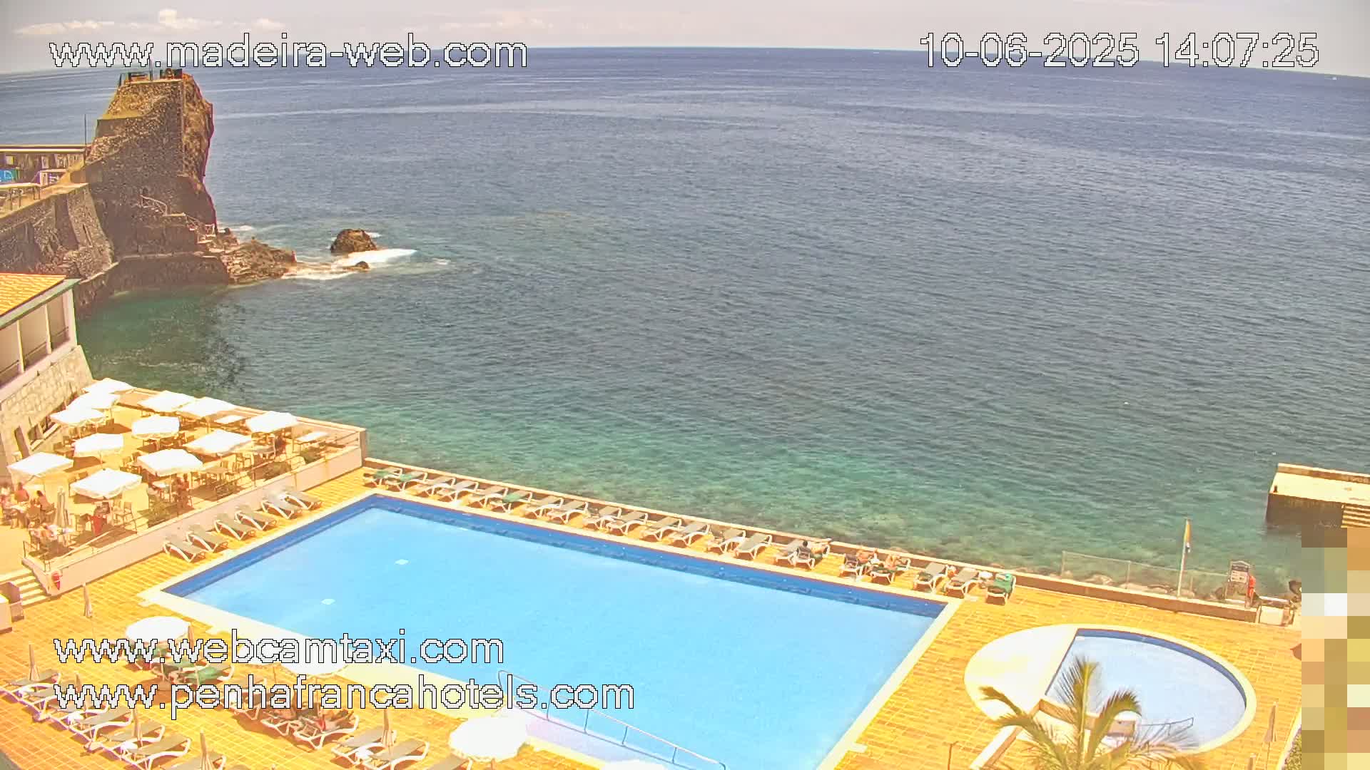 An outdoor swimming pool and sunbathing area overlook a calm ocean with a rocky outcrop in the distance under a sunny sky.