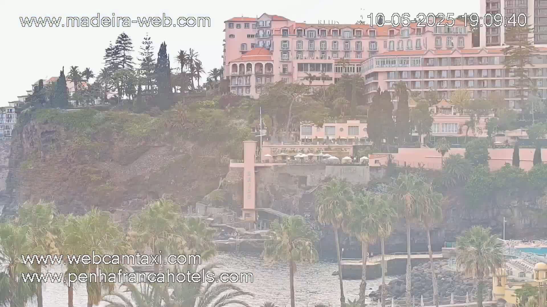 A large pink hotel sits atop a rocky cliff overlooking a calm ocean, surrounded by lush vegetation and palm trees under hazy skies.
