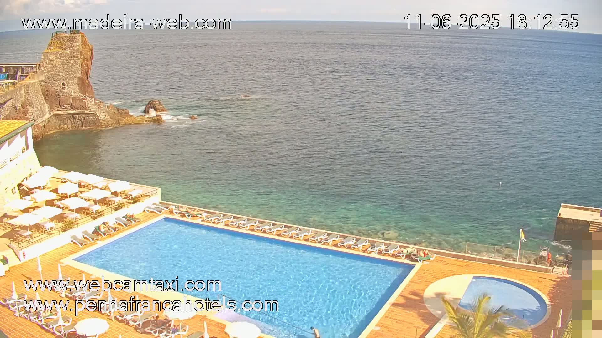 An aerial view shows a resort pool and patio area next to a rocky coast and calm ocean on a sunny day.