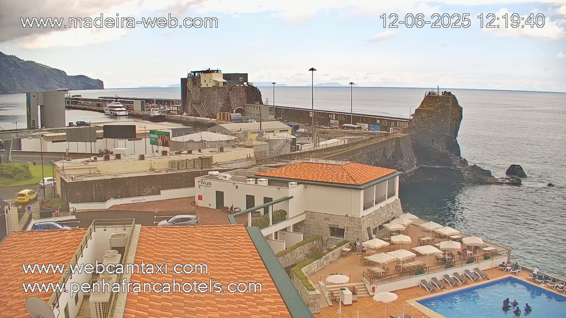 An overcast day shows a harbor with boats, buildings, a swimming pool, and patio furniture near a rocky coast.