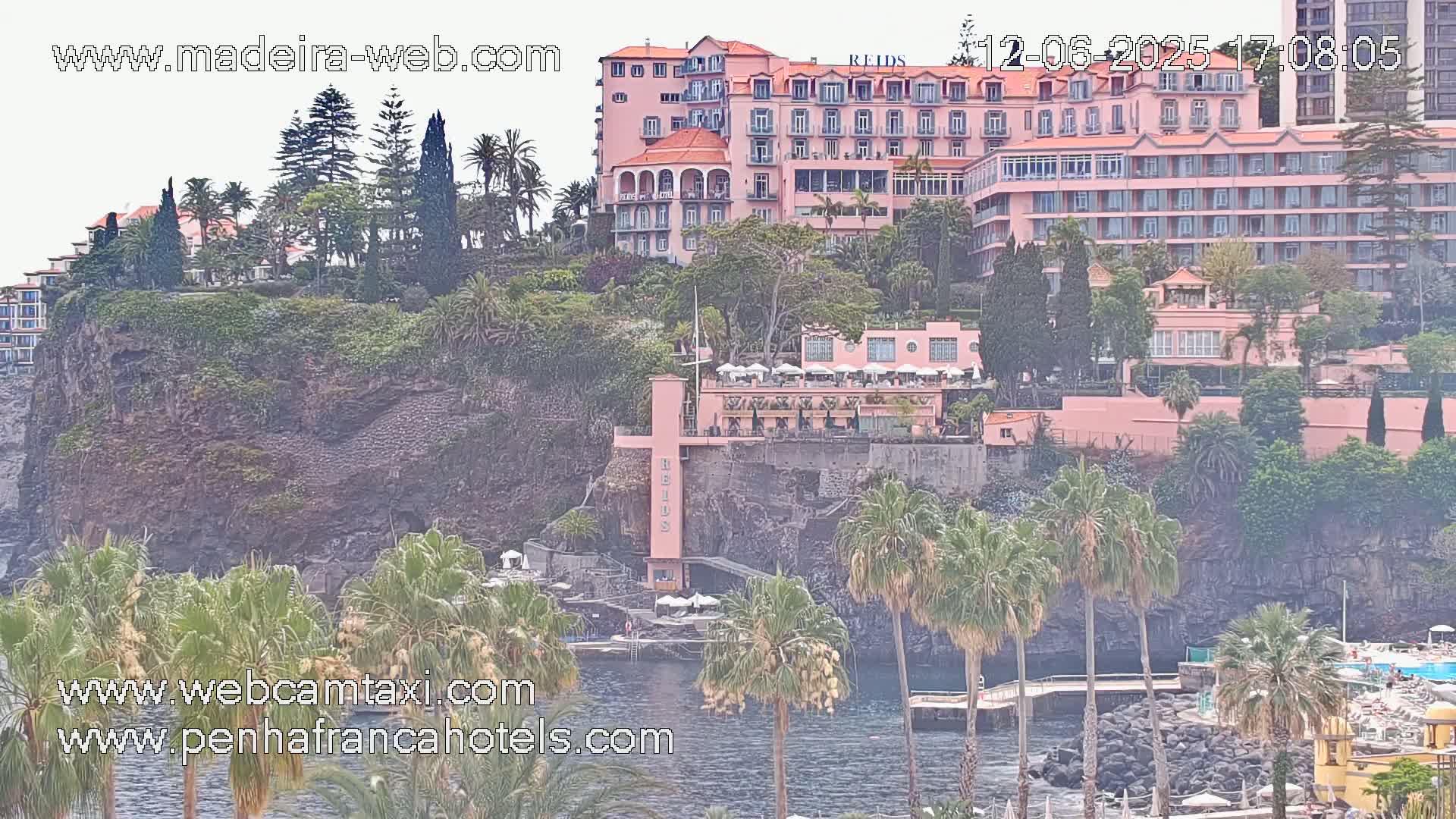 A large pink hotel complex is situated on a rocky cliff overlooking a calm body of water, surrounded by lush greenery and palm trees under an overcast sky.