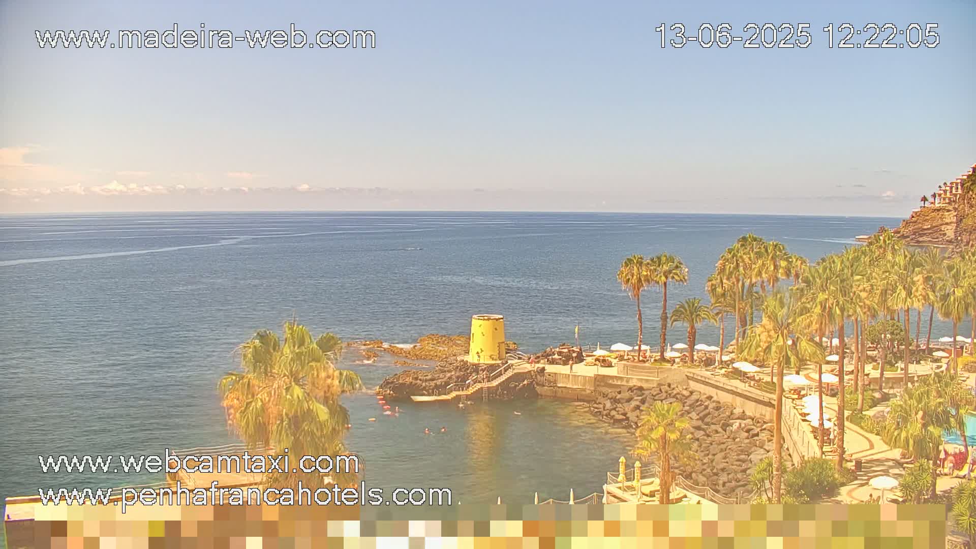 A clear, sunny day reveals a resort area with palm trees lining a rocky coastline and a calm ocean extending to the horizon.