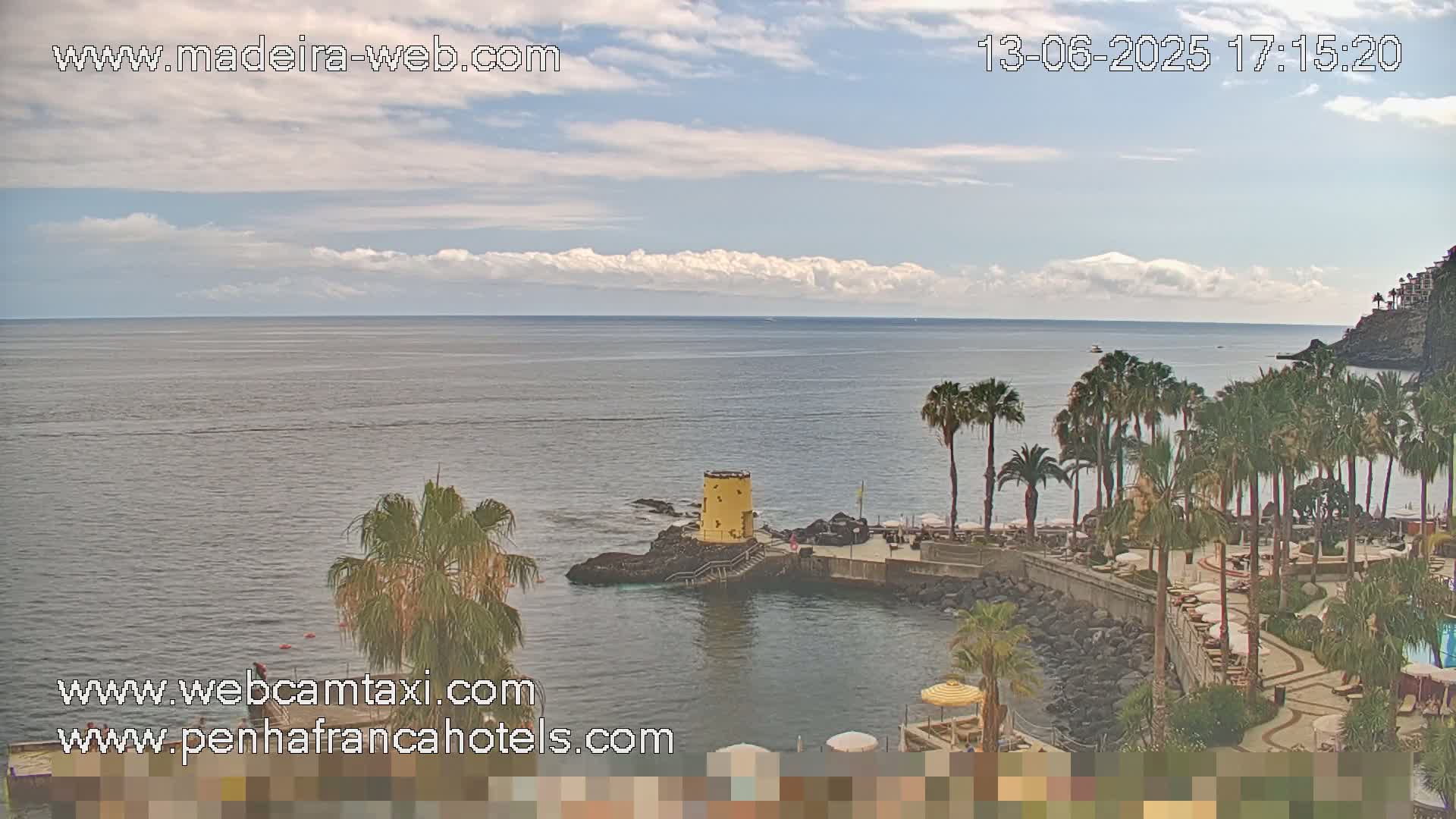 A partly cloudy day shows a calm ocean with a resort featuring palm trees, a yellow tower, and a rocky shoreline.