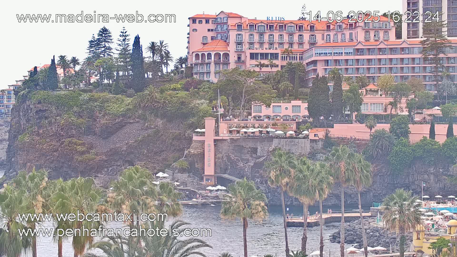 A large pink hotel complex is situated on a rocky cliff overlooking a calm body of water with palm trees and other vegetation surrounding it.