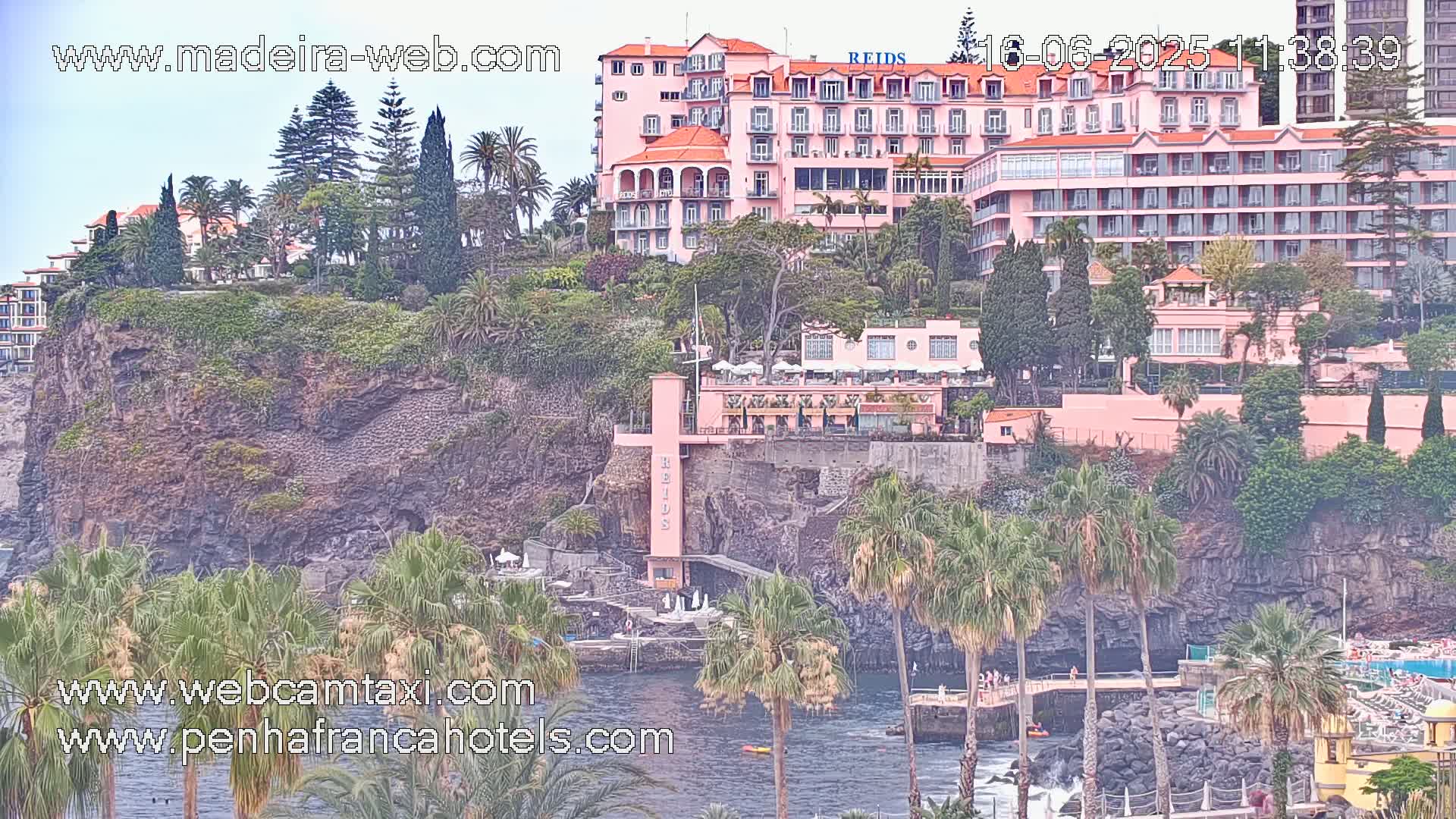 A pink hotel complex is perched on a rocky cliff overlooking a calm ocean, surrounded by lush tropical vegetation under a mostly clear sky.
