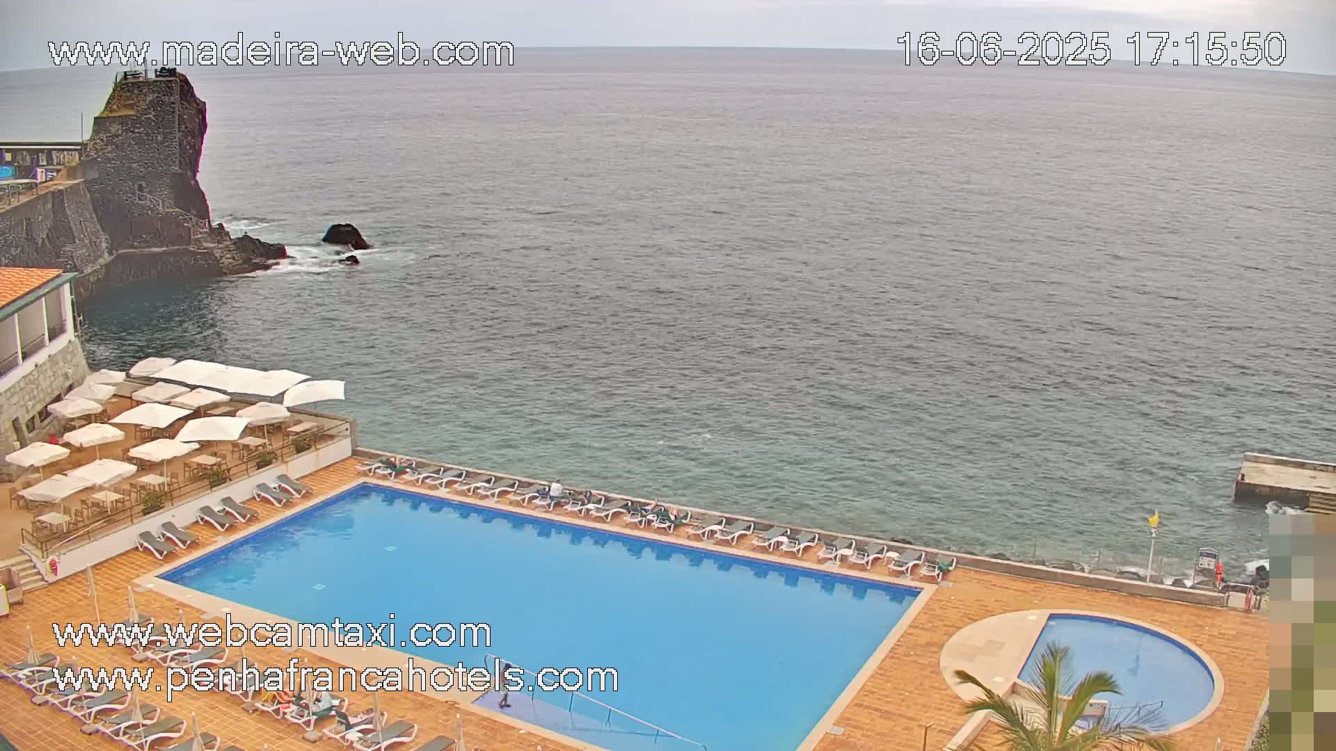 An outdoor swimming pool and lounge area at a hotel overlook a calm ocean on an overcast day.