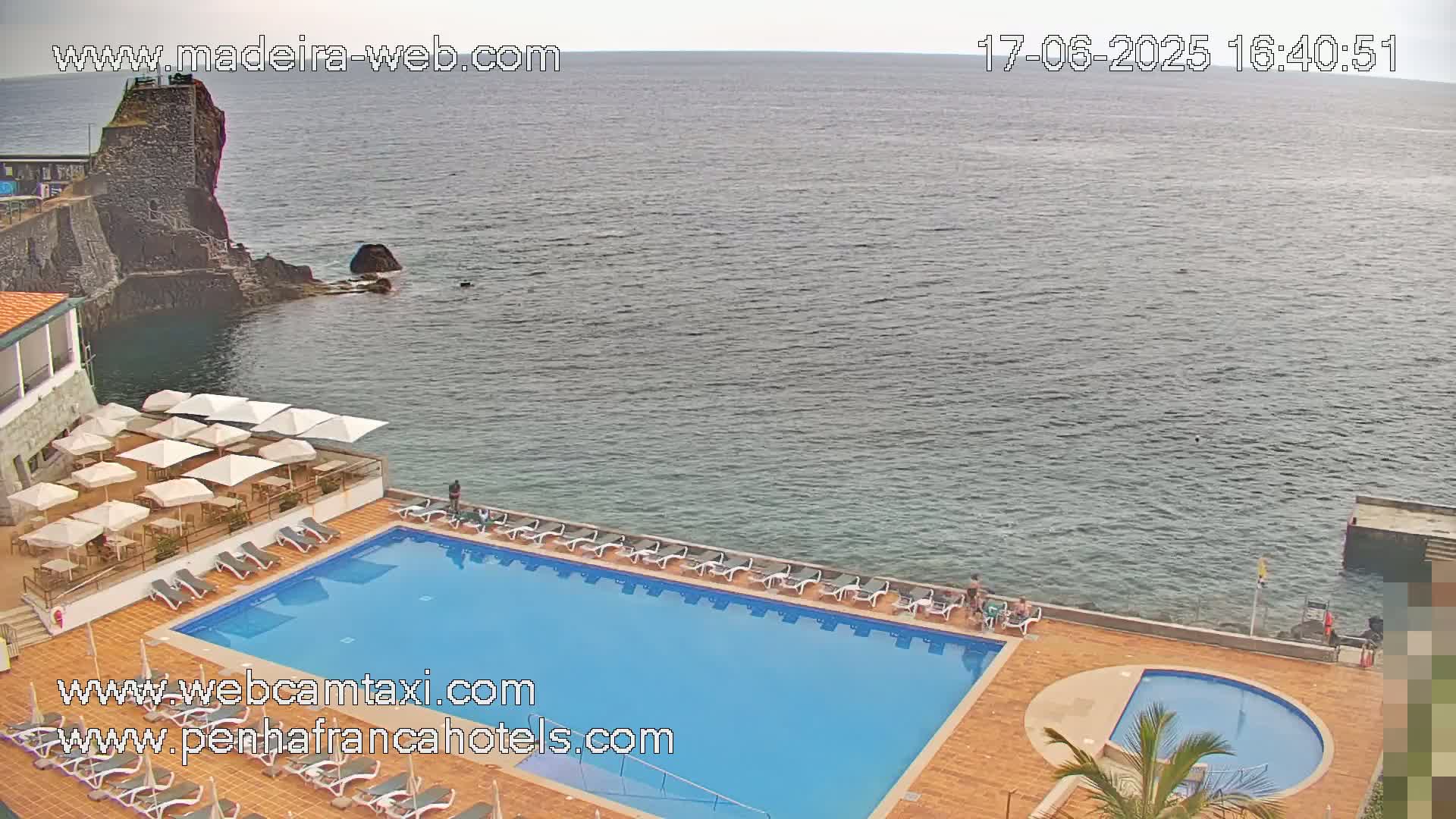 An outdoor pool and deck area with lounge chairs and umbrellas overlook a calm ocean next to a rocky cliff.