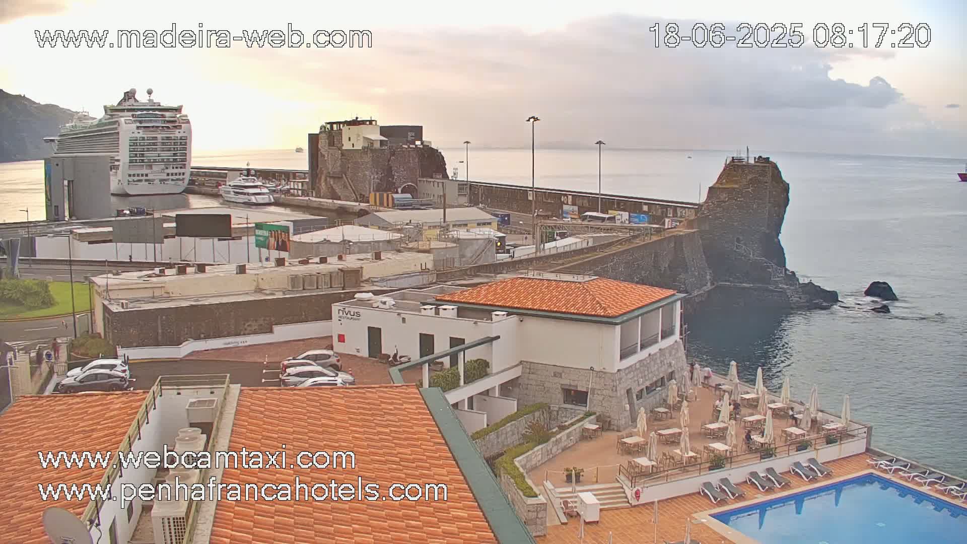 A cruise ship is docked at a harbor next to a rocky cliff, with a hotel and pool visible in the foreground, under partly cloudy skies.