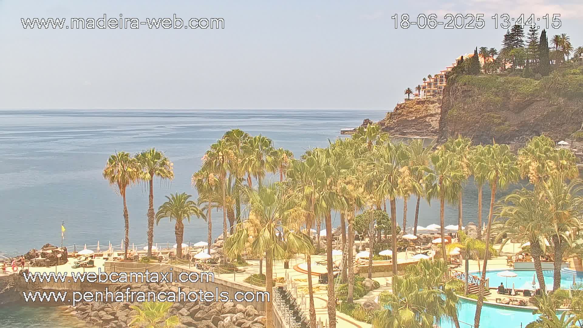 A resort with palm trees and pools overlooks a calm ocean and a clifftop building under a sunny sky.
