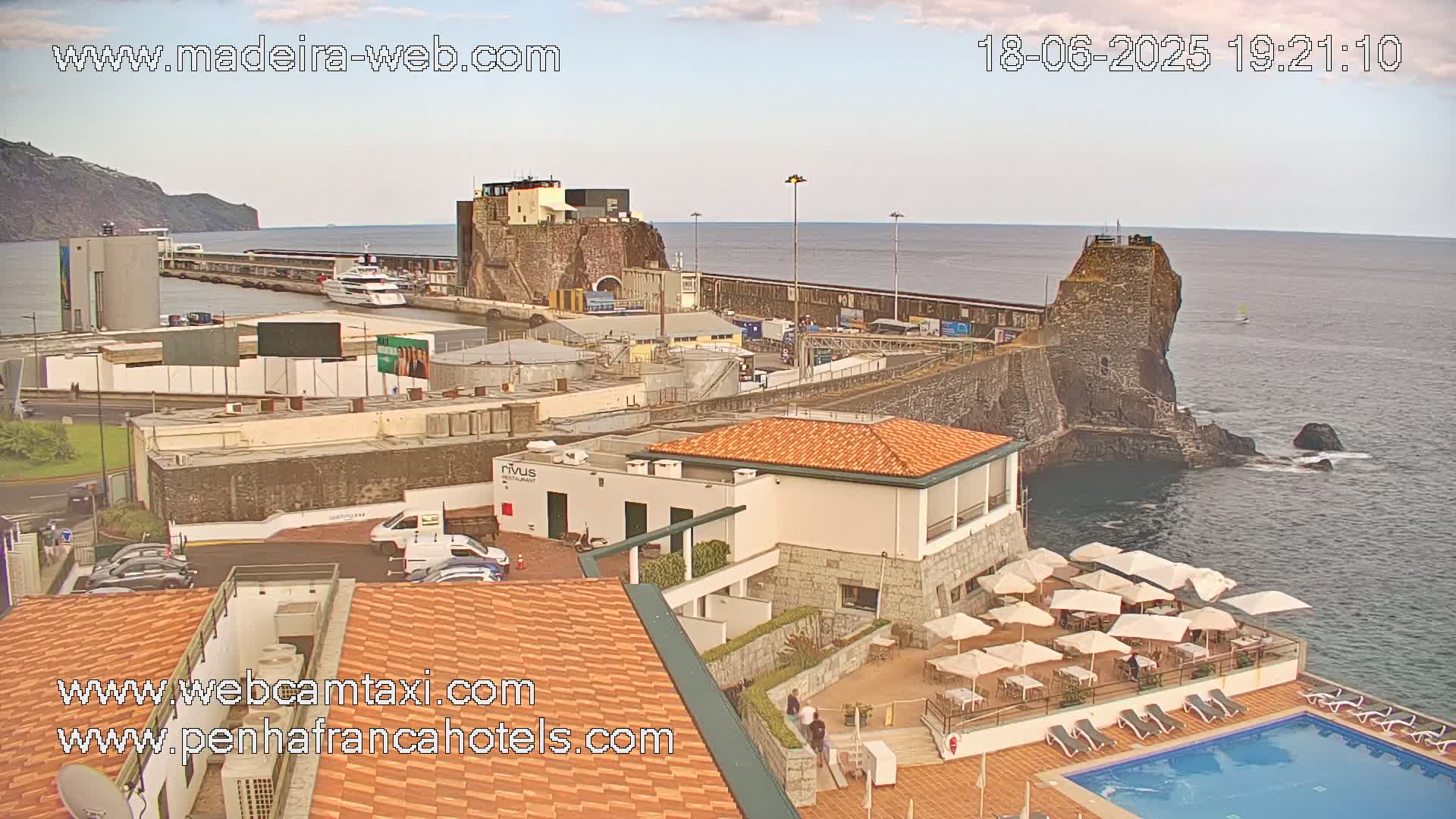 An oceanside resort with a swimming pool and outdoor seating overlooks a harbor with boats, a stone fortress, and a mountain in the background under partly cloudy skies.