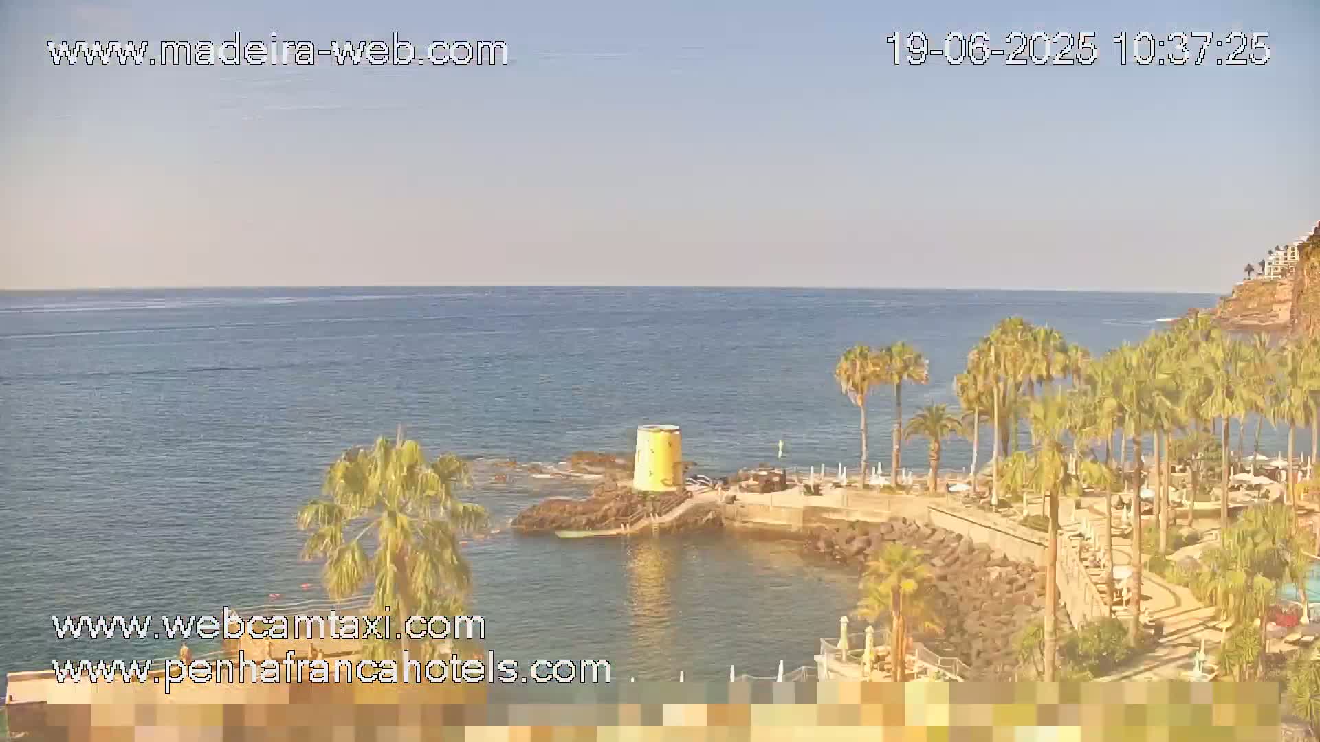 A clear, sunny day overlooking a calm ocean with palm trees lining a rocky coast and a small yellow structure near the water.
