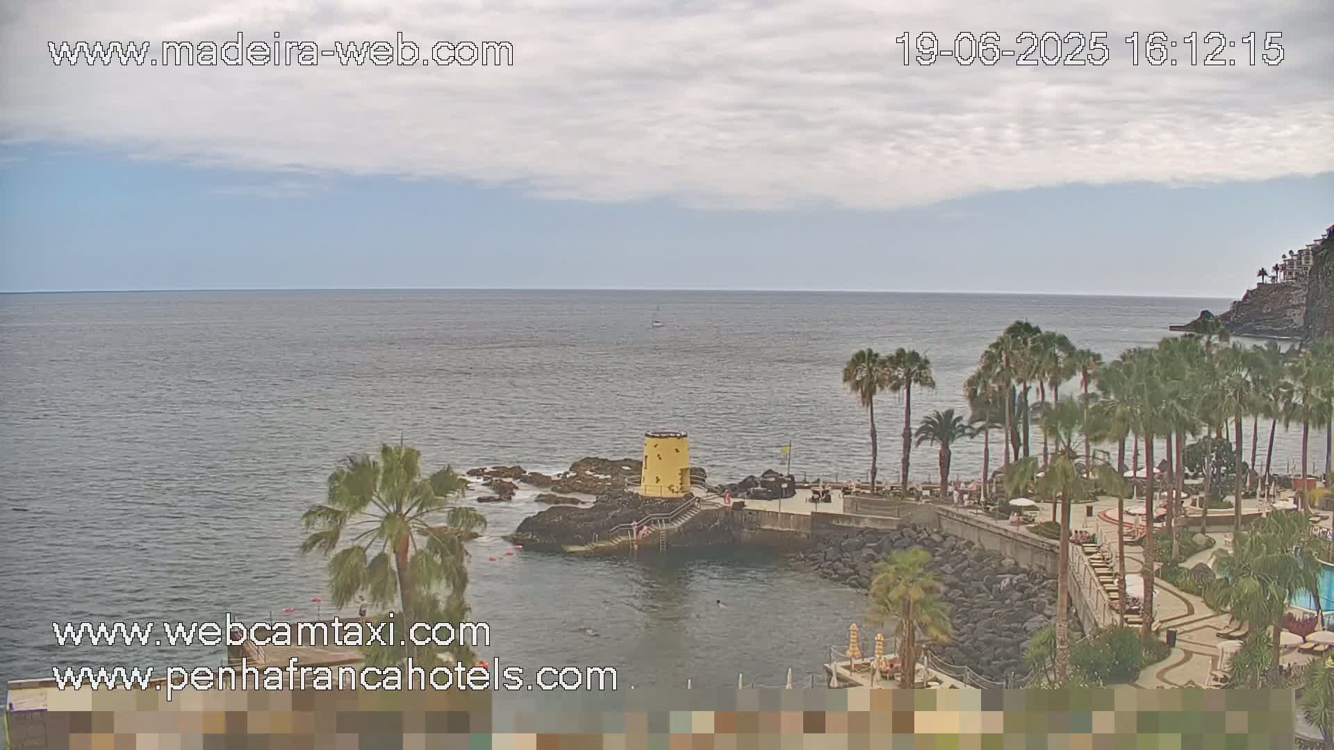 A partly cloudy day reveals a coastal scene with palm trees, a small yellow tower, a rocky shoreline, and calm ocean waters.