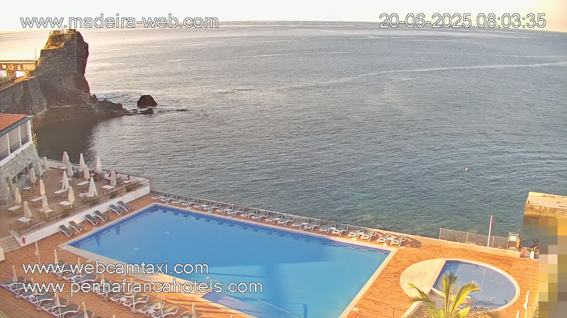 An outdoor pool with lounge chairs sits next to a rocky ocean shore under a clear sky.