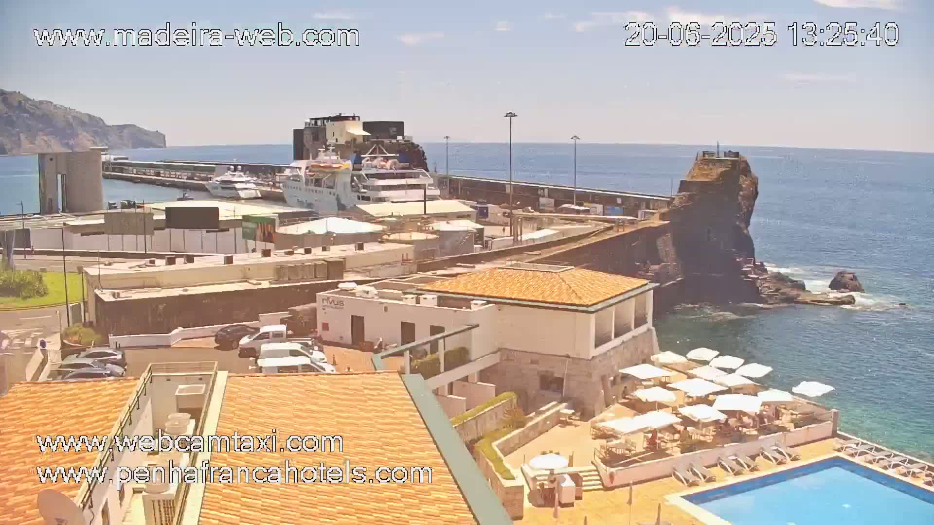 A sunny harbor scene shows docked boats, buildings, a pool with umbrellas and lounge chairs, and a rocky outcropping in the ocean.