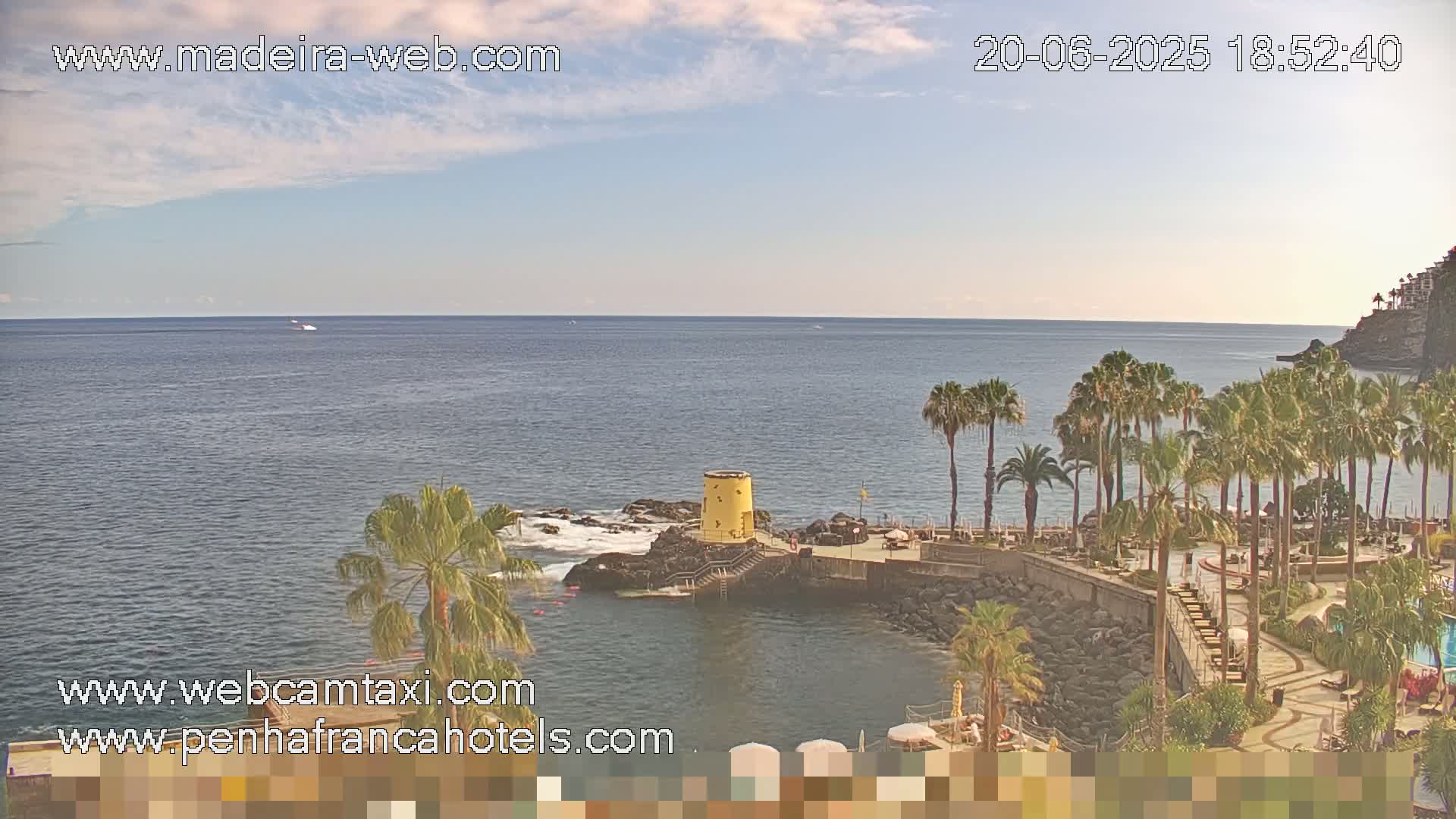 A partly cloudy day overlooks a calm ocean with palm trees and a resort by the water.