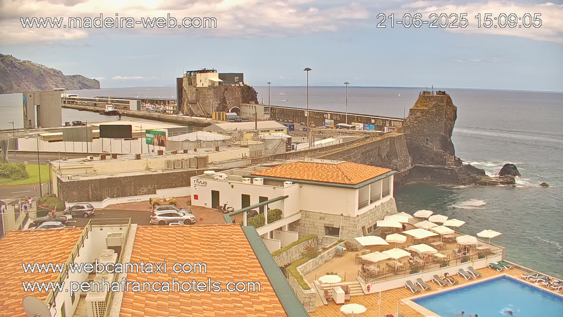 A coastal town with a harbor, buildings, and a hotel with a pool is seen under partly cloudy skies.