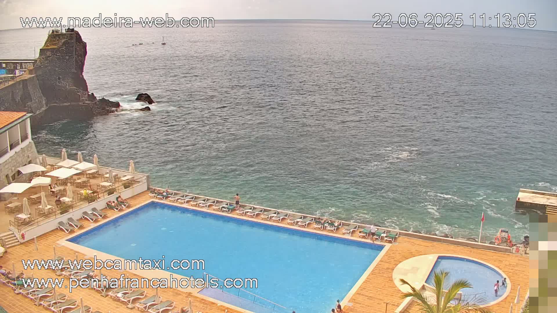 A resort pool with lounge chairs and a patio overlooks a calm ocean and dark rocky shoreline.