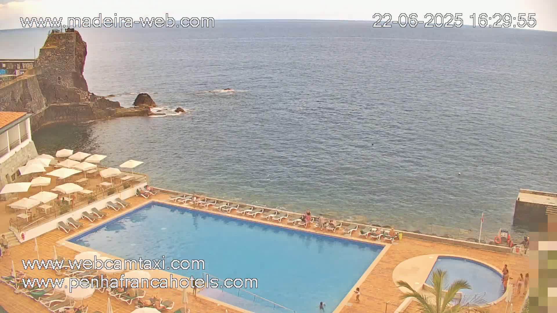An outdoor swimming pool with surrounding lounge chairs and umbrellas is situated next to a calm ocean, with a rocky cliff and old stone structure visible in the distance.