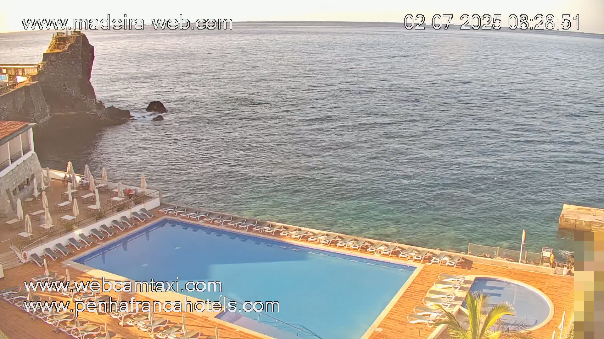 An outdoor swimming pool with lounge chairs is situated next to a rocky ocean shore under a mostly clear sky.