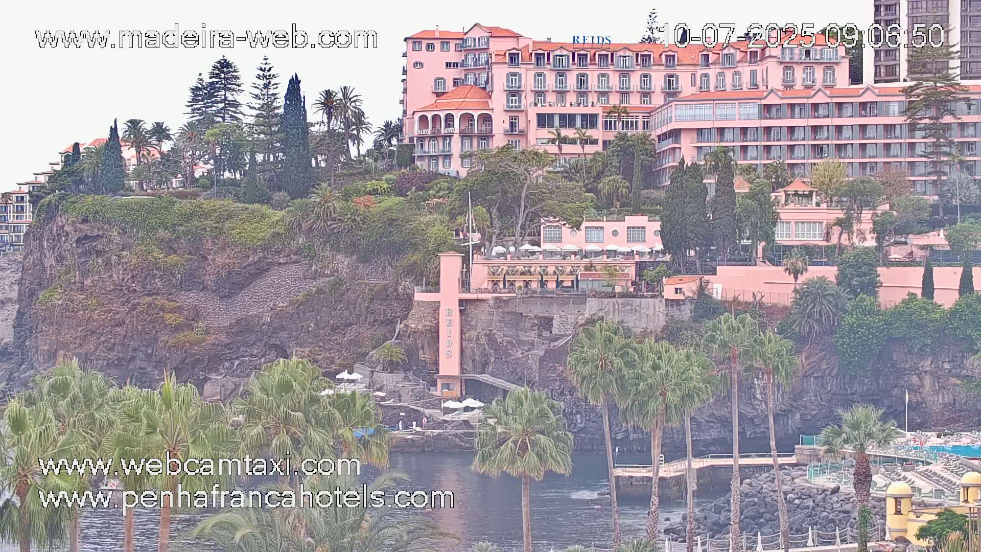A large pink hotel is situated on a rocky cliff overlooking a calm body of water, surrounded by lush vegetation including palm trees under an overcast sky.