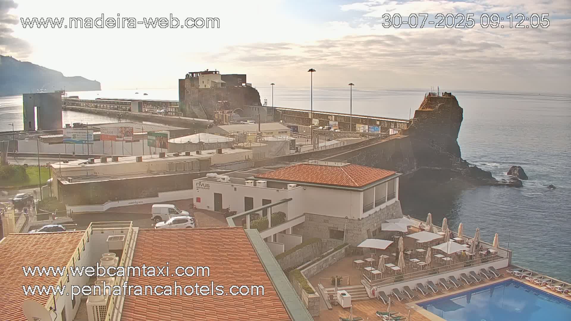 An oceanside view shows a coastal town with buildings, a harbor, a rocky outcropping, a swimming pool, and a mostly clear sky.